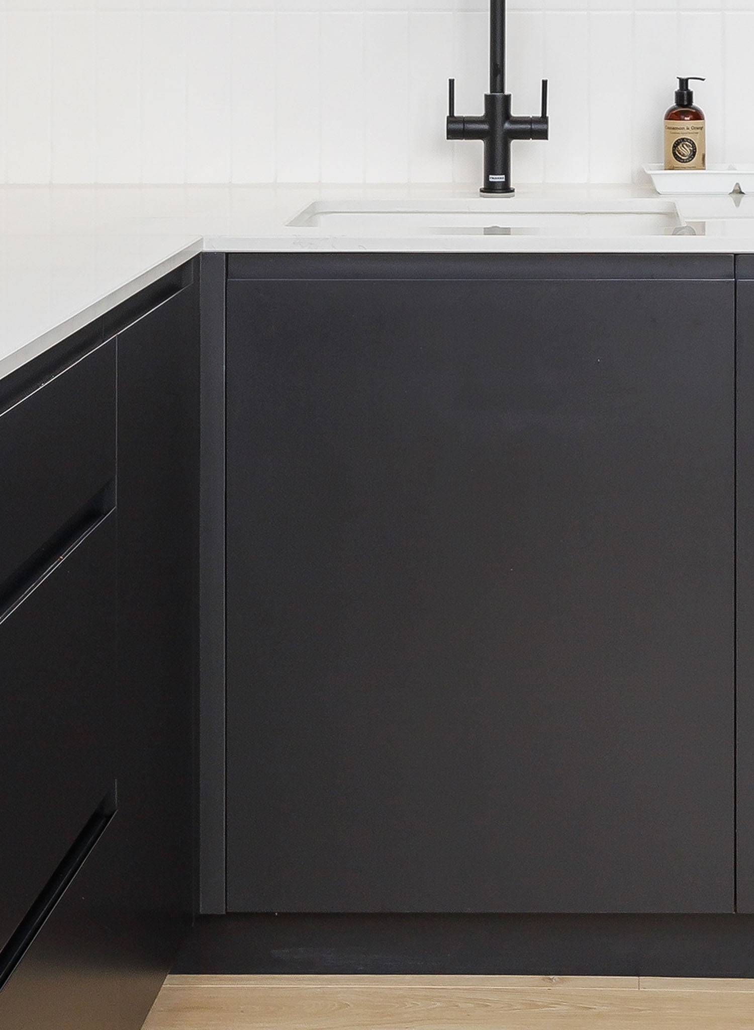Modern kitchen with matte black cabinets, white countertop, black faucet, built-in sink, and a soap dispenser on the counter. Light wood flooring is partially visible at the bottom.