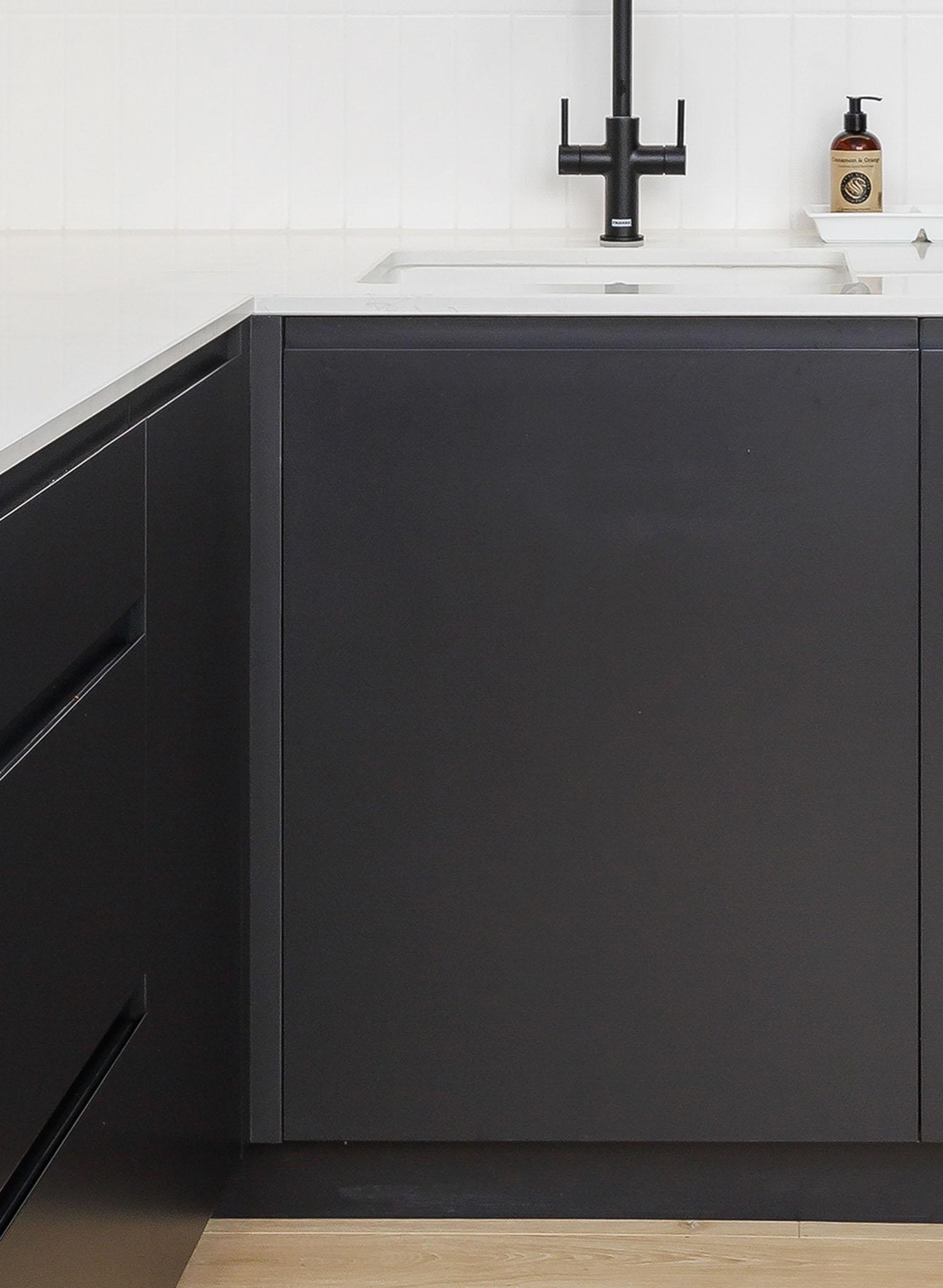 Modern kitchen with matte black cabinets, white countertop, black faucet, built-in sink, and a soap dispenser on the counter. Light wood flooring is partially visible at the bottom.