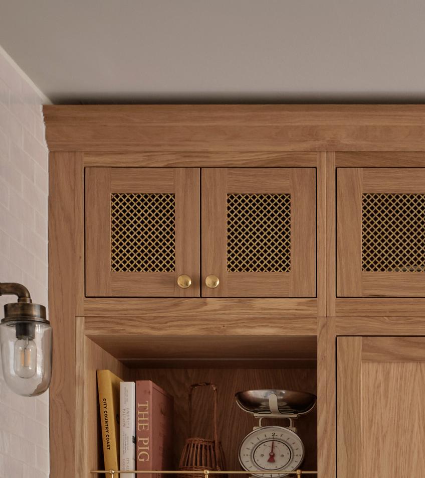 A wooden kitchen cabinet with mesh doors and gold knobs. Below, a shelf holds books, a decorative bowl, and a vintage kitchen scale. A wall sconce with a glass shade is mounted on the left.