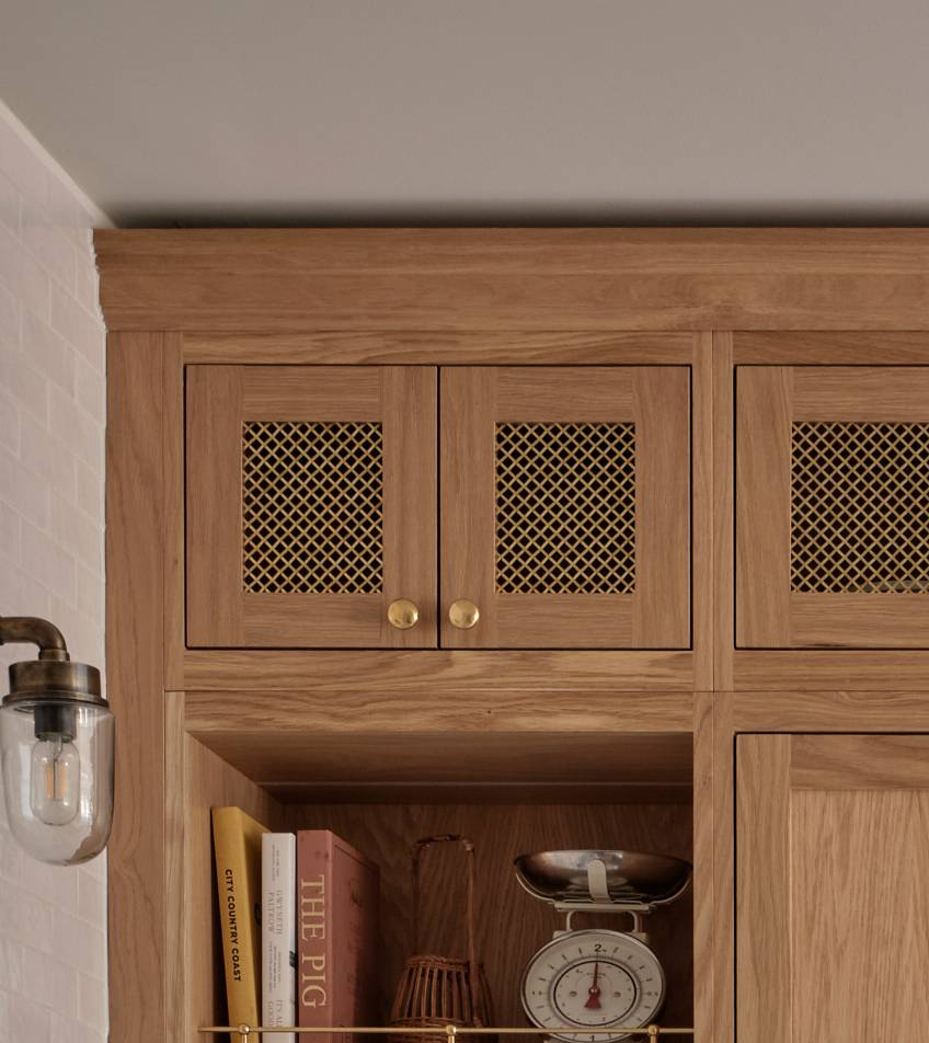 A wooden kitchen cabinet with mesh doors and gold knobs. Below, a shelf holds books, a decorative bowl, and a vintage kitchen scale. A wall sconce with a glass shade is mounted on the left.