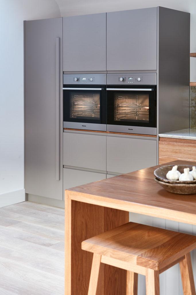 A modern kitchen setup featuring a grey floor-to-ceiling cabinet unit with two integrated stainless steel ovens side by side, with a wooden dining counter and stool in the foreground.