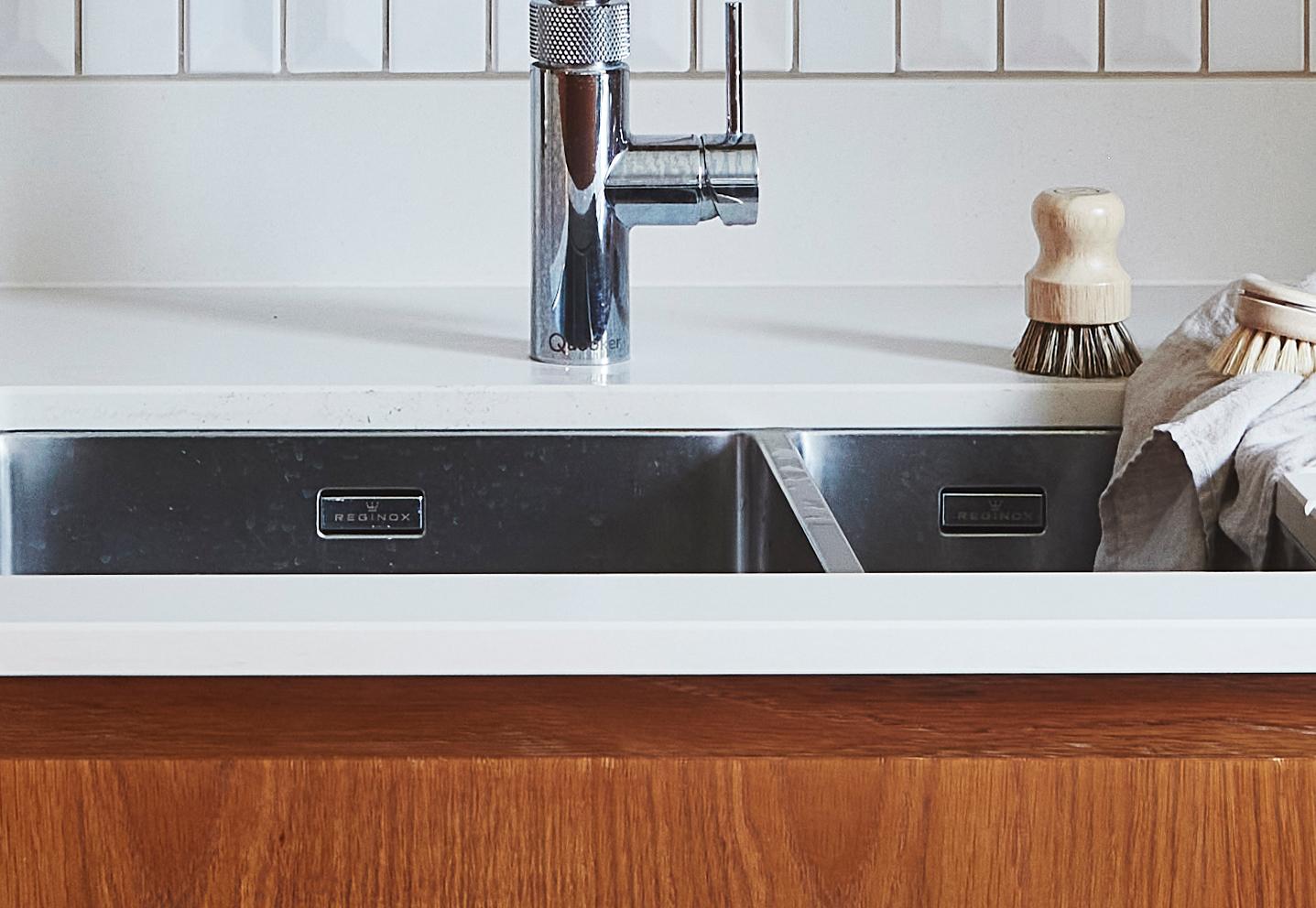 A modern kitchen sink with a stainless steel faucet, white countertop, and wooden cabinet. A dish brush with a wooden handle stands beside a dishcloth draped over the side of the sink. White tile backsplash in the background.
