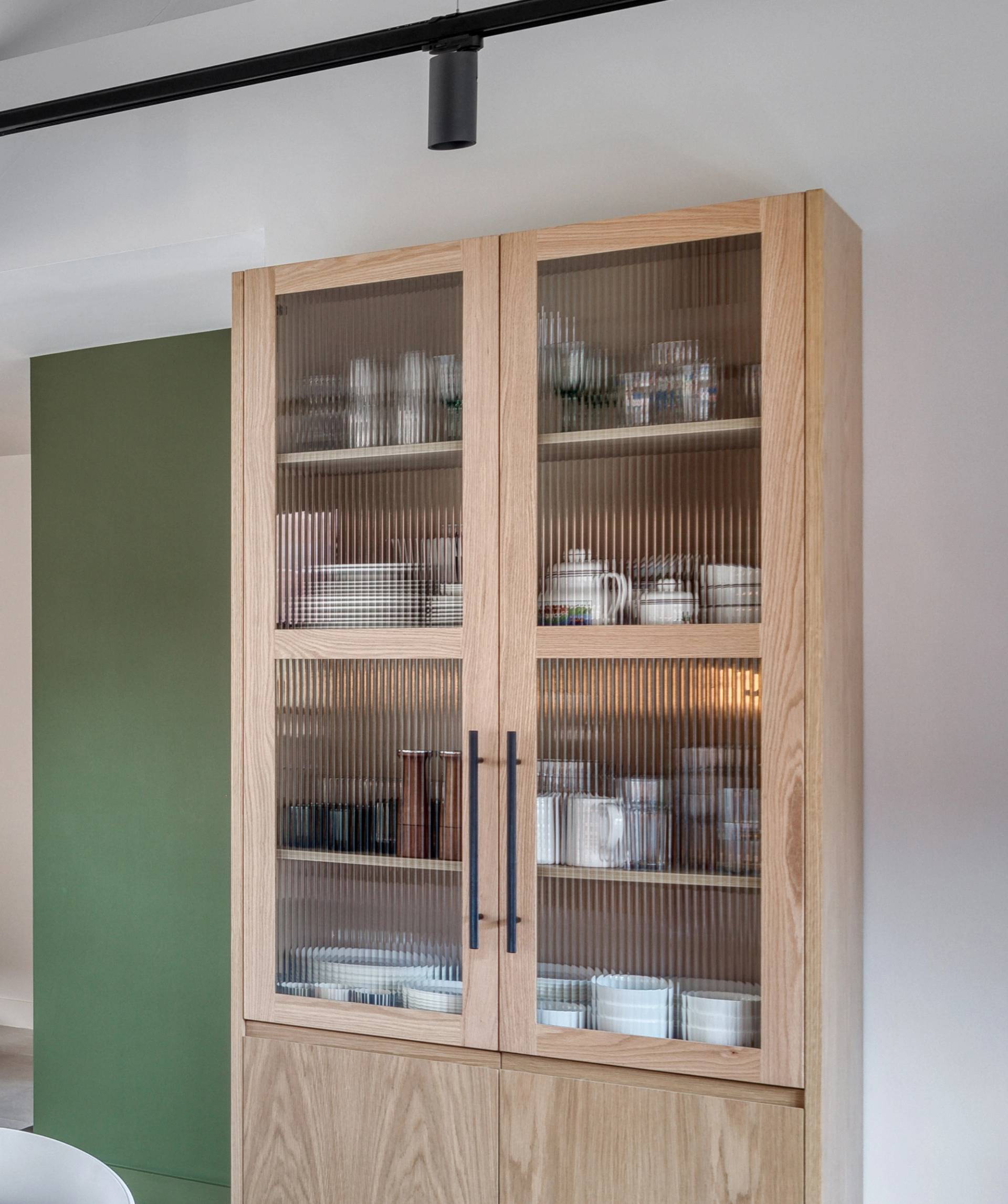 A wooden kitchen cabinet with reeded glass doors displays neatly arranged dishes, cups, and bowls. The cabinet is situated against a wall with a green accent panel. Overhead track lighting is visible.