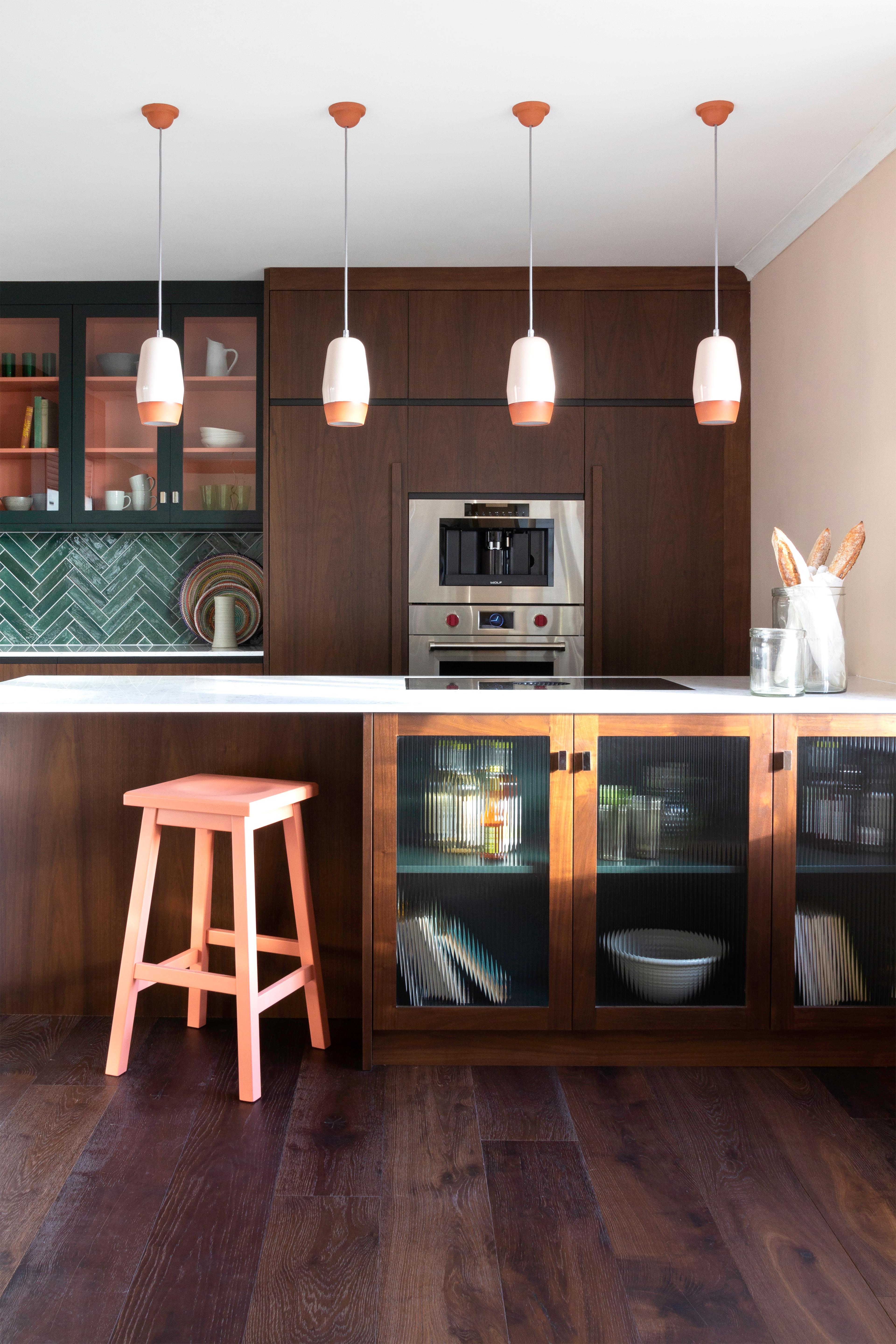 a vibrant kitchen features a dark wood island with ribbed glass doors, a single coral-colored bar stool, matching terracotta-accented pendant lights, and integrated appliances set against dark wood cabinetry.