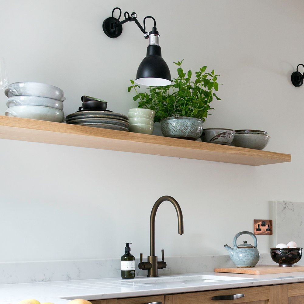 A minimalist kitchen features a wooden shelf holding stacked plates, bowls, cups, and a potted herb under a black wall lamp. Below, a sink with a gold faucet, soap dispenser, and teapot are set on a white countertop.