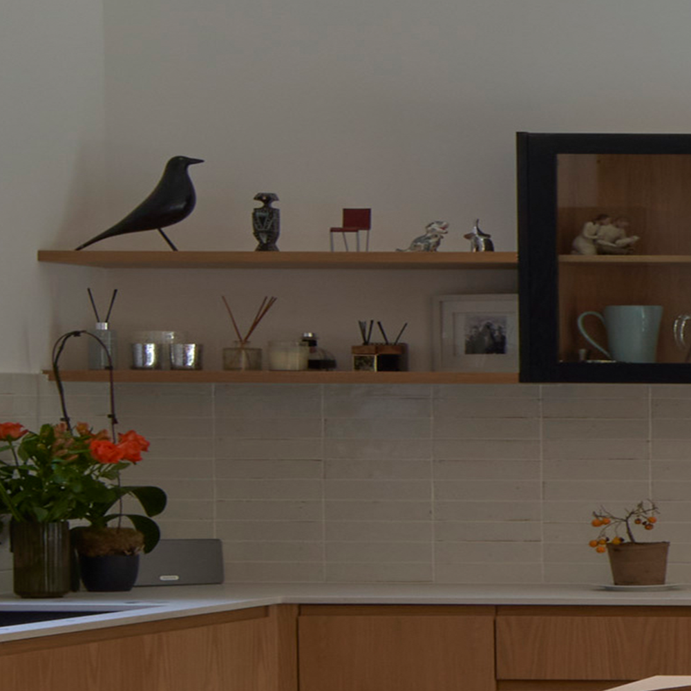 A modern kitchen corner with wooden cabinets, wall shelves holding decorative items, candles, a black bird sculpture, and potted orange flowers on the counter. A light tiled backsplash is visible.