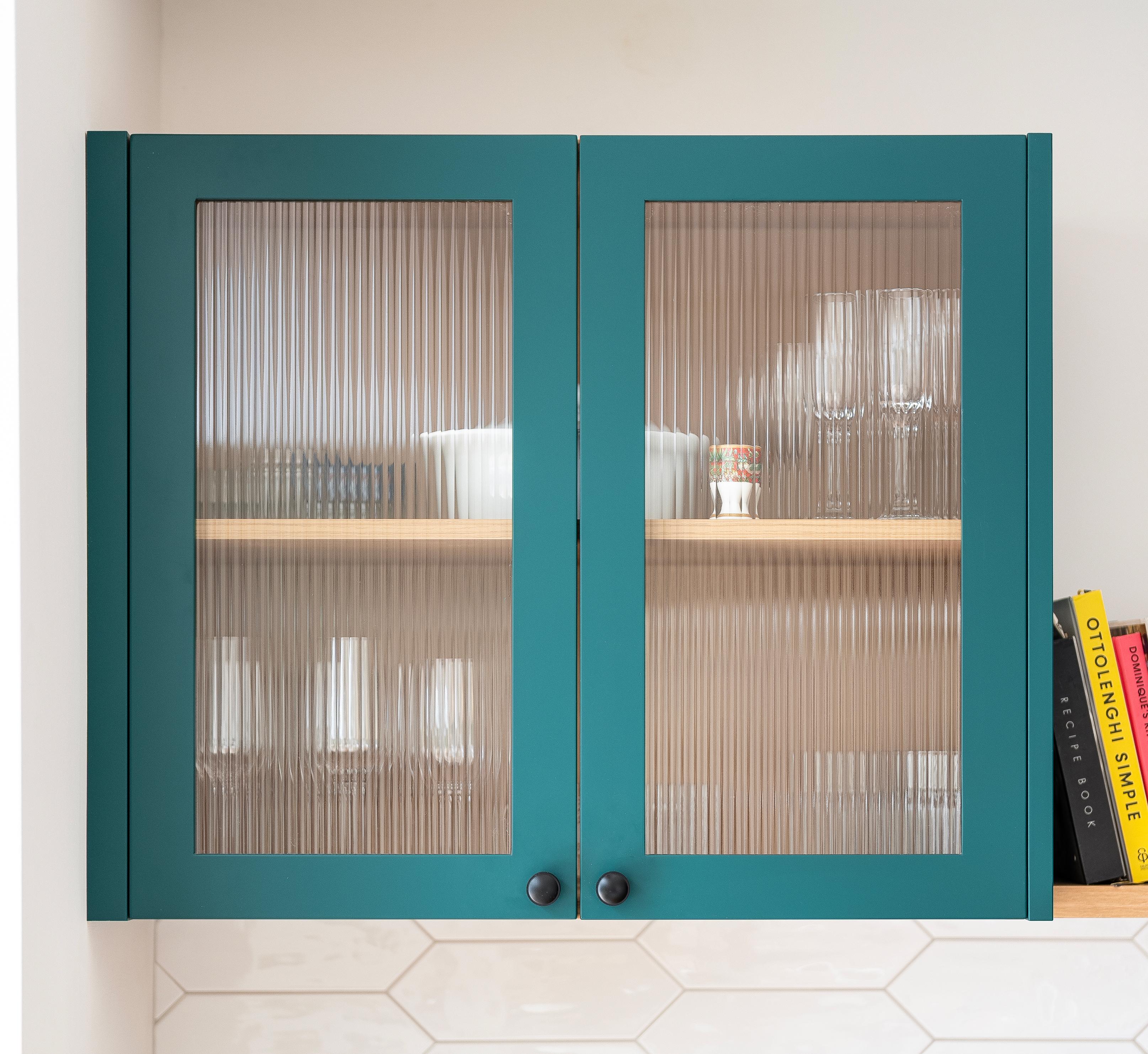 A teal kitchen cabinet with ribbed glass doors displays glassware and dishes on wooden shelves. A few books are stacked vertically on an adjacent shelf to the right. White hexagonal tiles line the wall below.