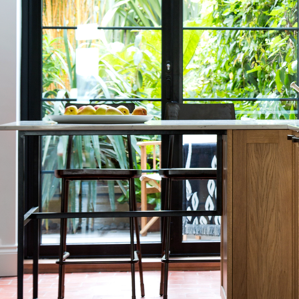 A modern kitchen with a wooden island, two barstools, and a bowl of assorted fruit. Large glass doors in the background reveal lush greenery outside.