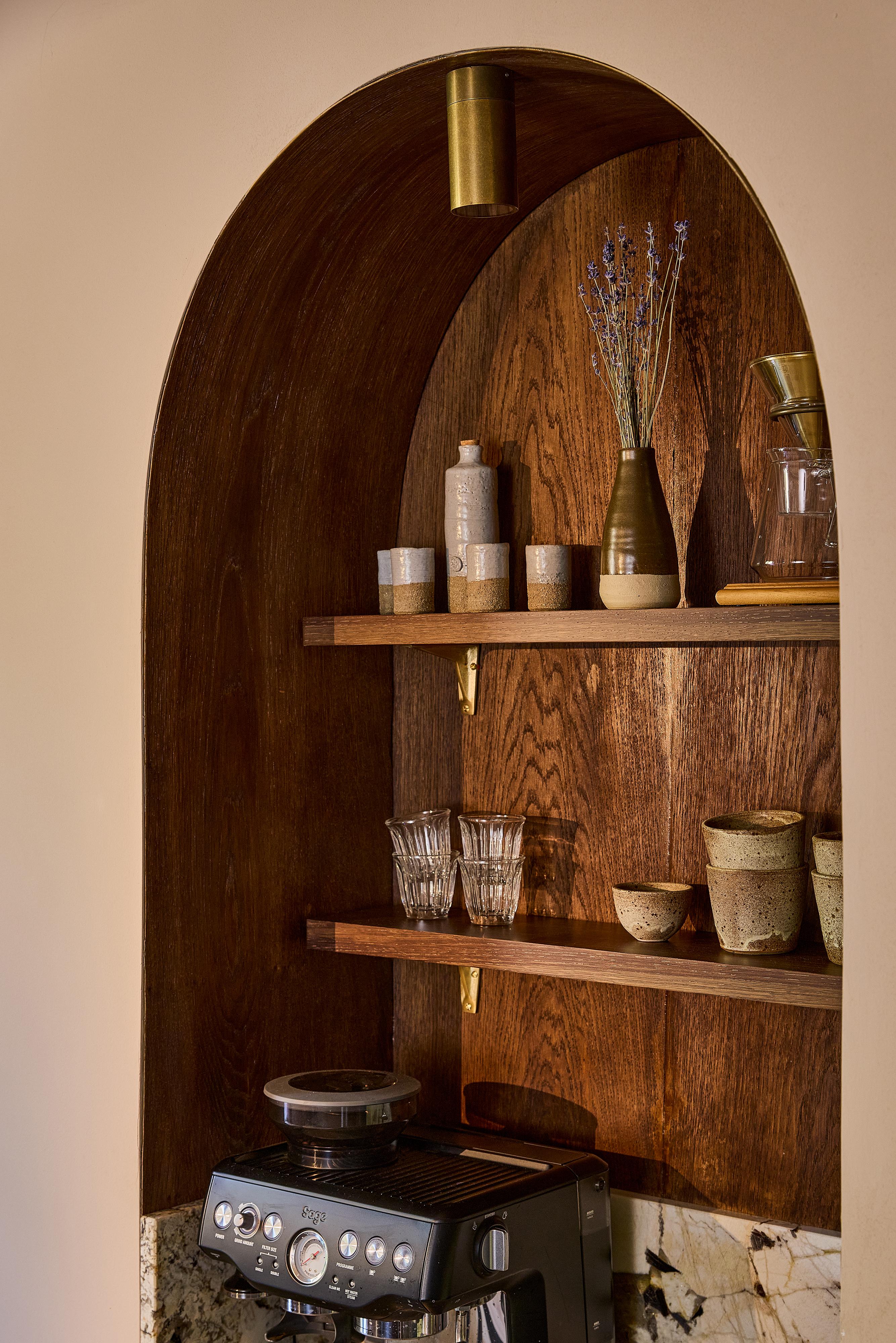 A wooden alcove with shelves holds ceramic cups, bowls, glasses, a vase with dried lavender, and a coffee maker below. The background is warm-toned with a marble counter edge visible at the bottom.
