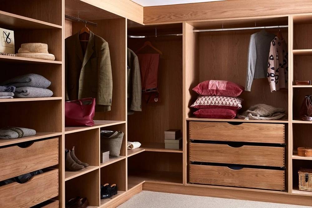 A wooden wardrobe with open shelves and hanging space. It contains folded clothes, a handbag, shoes, and a few hanging jackets and a sweater. There are pillows on one shelf and two drawers underneath with more folded items.