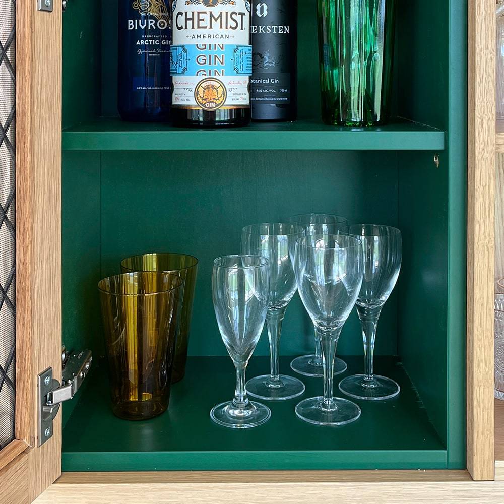 A green cabinet with bottles of spirits on the top shelf and a lower shelf holding four brown tumblers and six clear wine glasses. The cabinet door is partly open, showing a wooden frame and metal hinge.