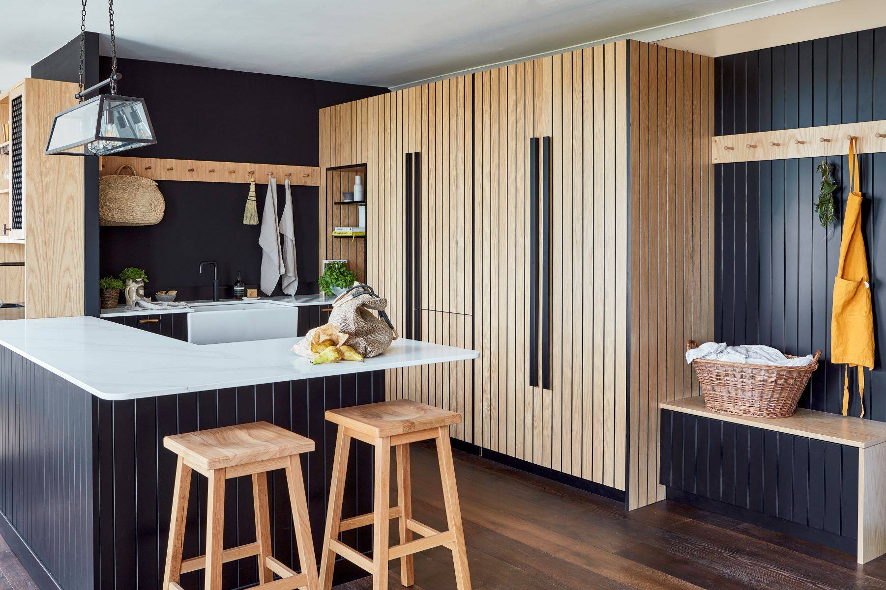 dark blue cabinets with white worktop in L shape. Tall oak cabinets with black vertical grooves. Bar handles on doors.