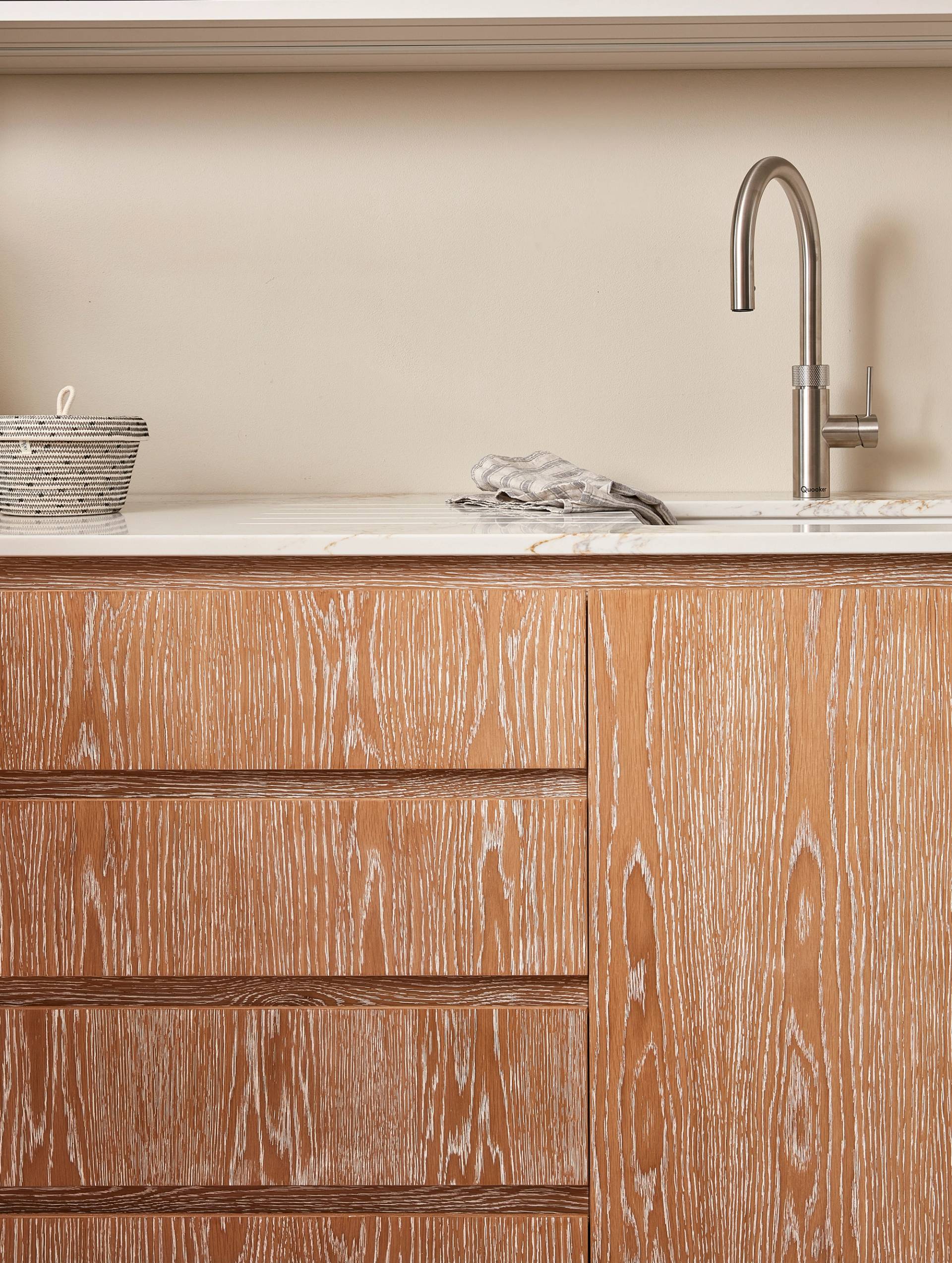 A wooden kitchen cabinet with a whitewashed finish under a countertop. A modern stainless steel faucet is mounted on the countertop beside a small basket. A soft cloth is placed next to the sink, and the wall is a light cream color.