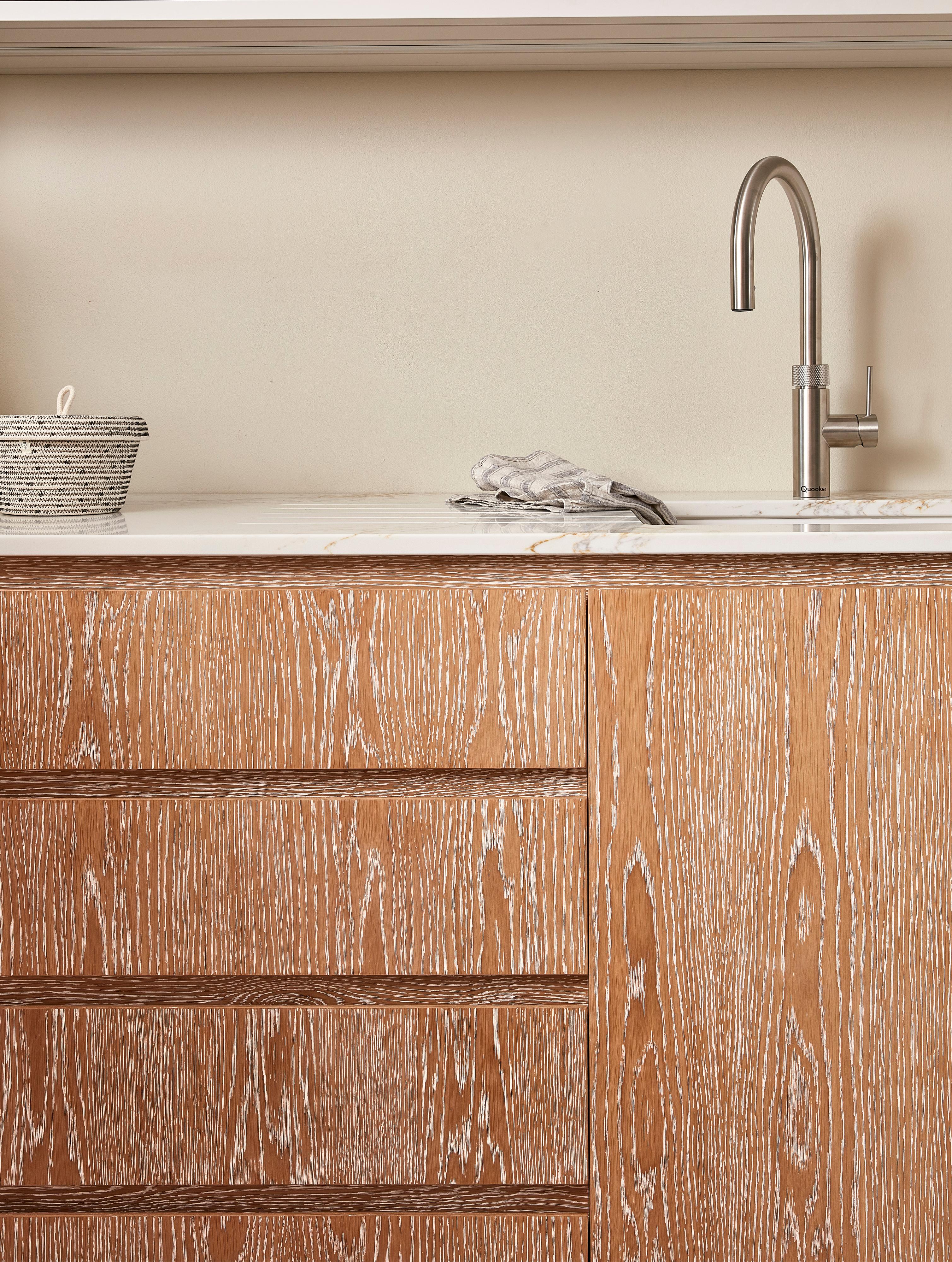 A wooden kitchen cabinet with a whitewashed finish under a countertop. A modern stainless steel faucet is mounted on the countertop beside a small basket. A soft cloth is placed next to the sink, and the wall is a light cream color.