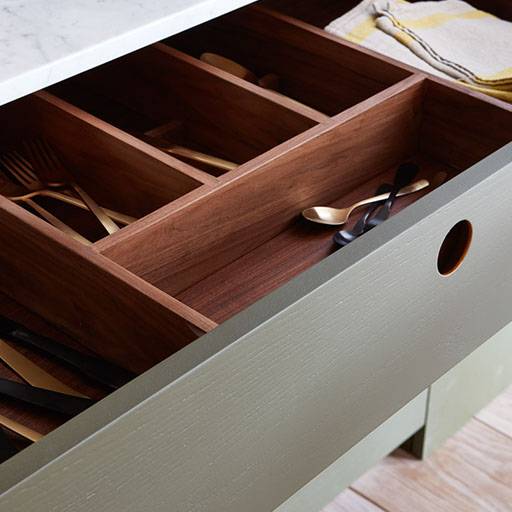 An open kitchen drawer with a wooden organizer holding gold and black cutlery, including spoons and forks. A yellow and white towel is partially visible on the countertop above the drawer.
