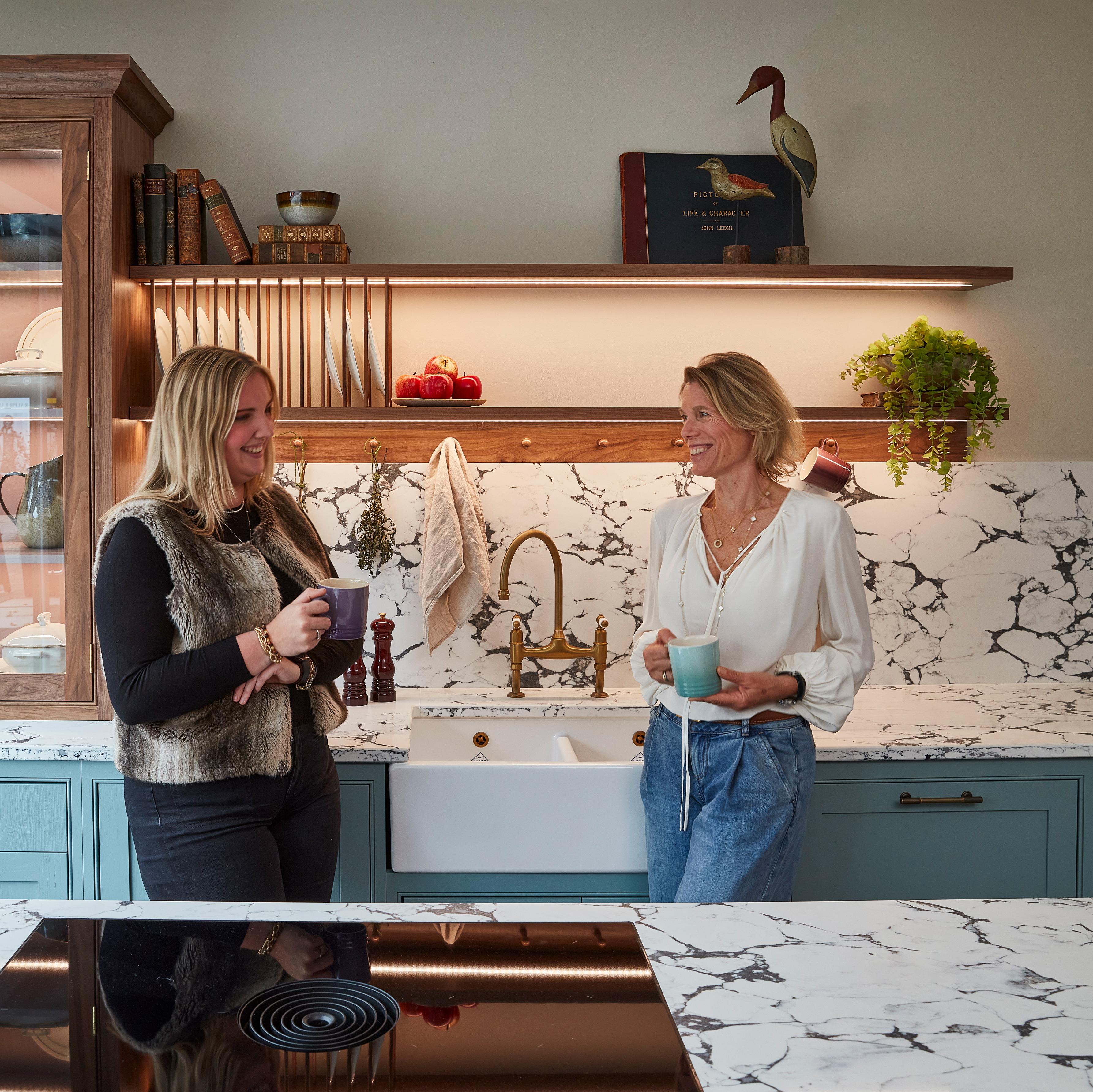 Two women smiling and talking in a modern, luxury kitchen while holding coffee mugs. The kitchen features marble countertops, a white farmhouse sink, and teal-colored cabinetry with warm under-shelf lighting.