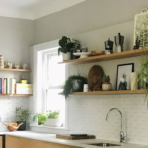 A modern kitchen with open wooden shelves holding plants, books, dishes, and decor above a white countertop and sink, set against a white tile backsplash and a window letting in natural light.