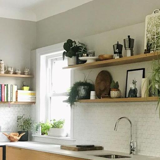 A modern kitchen with open wooden shelves holding plants, books, dishes, and decor above a white countertop and sink, set against a white tile backsplash and a window letting in natural light.