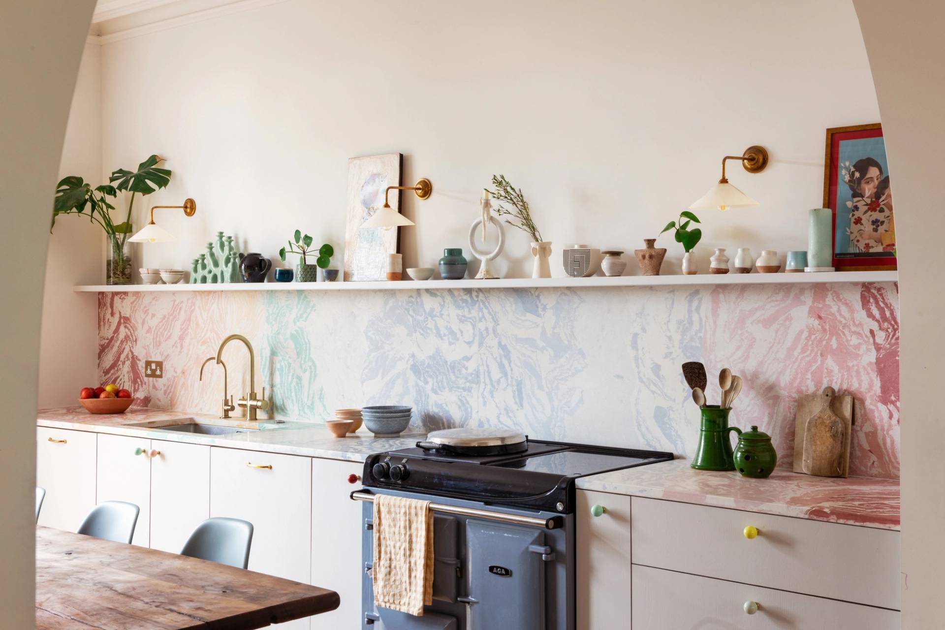A cozy kitchen with a marble-patterned backsplash, white cabinets, a brass faucet, open shelves holding plants, dishes, and art, and a blue stove with a yellow towel. Warm lighting and soft colors create an inviting atmosphere.
