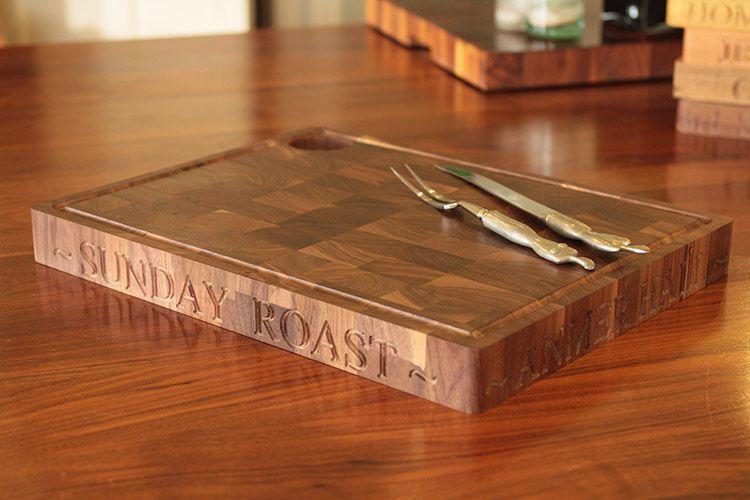 Wooden chopping board engraved with 'Sunday roast' set on a wooden table. Two carving utensils, knife and fork with metallic handles, are placed on the board