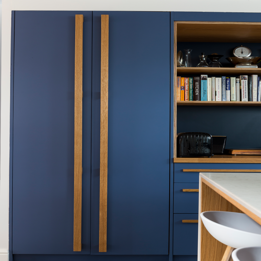 A modern kitchen with matte blue cabinets featuring vertical wooden handles, open shelving with books and kitchen items, a black toaster, and a white countertop with a white bar stool in the foreground.