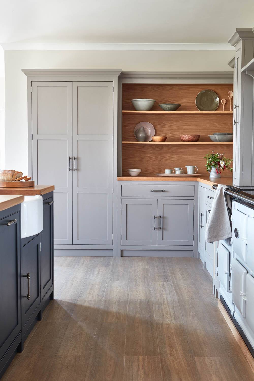 A modern kitchen with light gray cabinets, a wooden countertop, and open shelves displaying dishes and bowls. The floor has wood-look tiles, and a large farmhouse sink is visible on the left. Natural light brightens the space.
