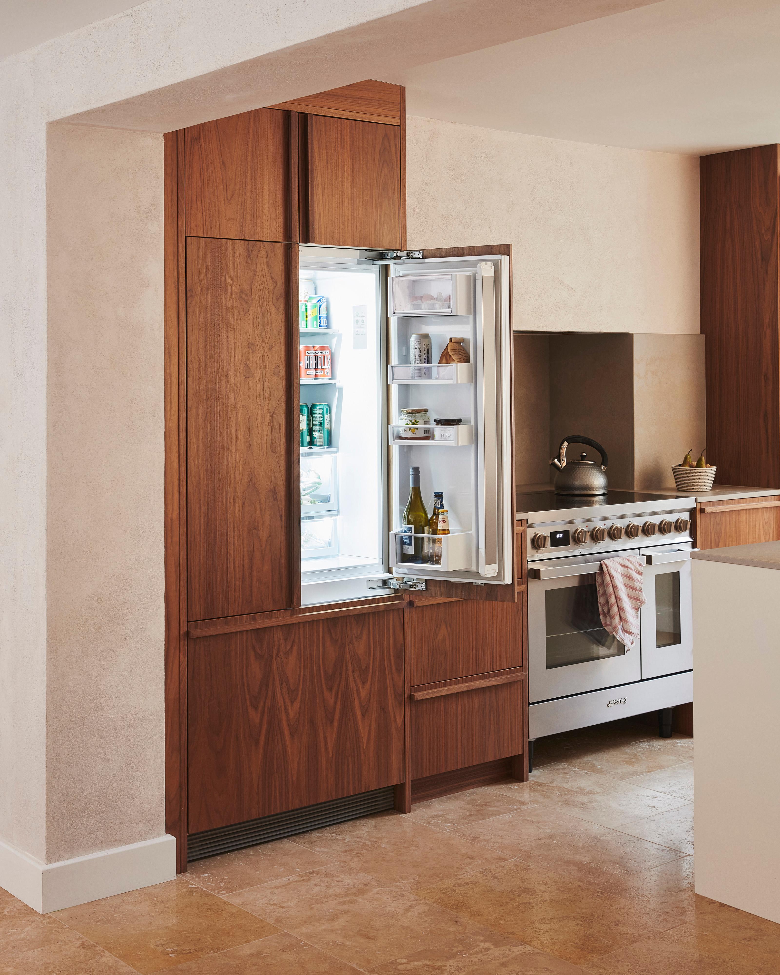 A modern kitchen with wooden cabinets, a stainless steel oven, and a white countertop. A kettle sits on the stovetop, and a small plant is placed on the counter next to the oven. The floor is made of light beige tiles.