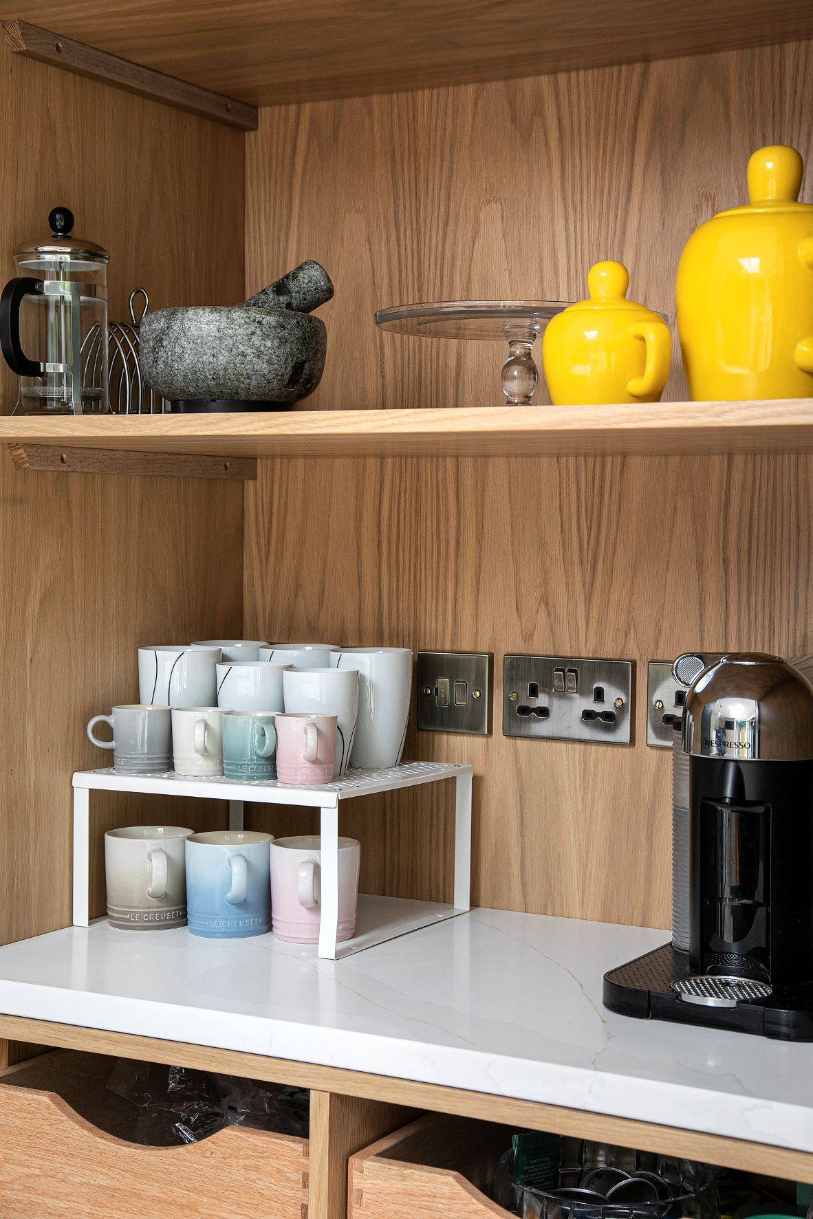 A wooden kitchen shelf with a mortar and pestle, a French press, yellow teapots, mugs in pastel colors on a white rack, pitchers, a coffee machine, and multiple power outlets on a white countertop.