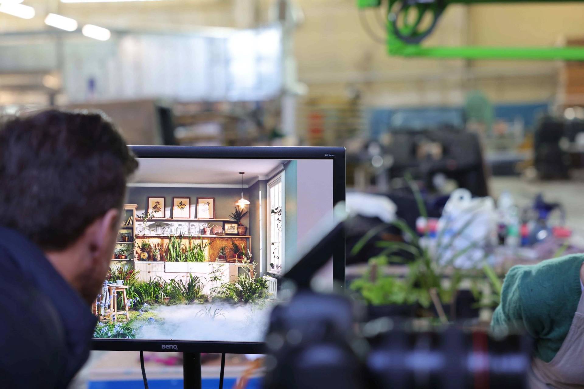 A behind-the-scenes shot of a professional production set, focusing on a BenQ monitor displaying a high-resolution image of a surreal, plant-filled kitchen. On the screen, the kitchen is seen with low-lying mist or fog rolling over a mossy floor. The back of a person's head is visible in the foreground as they review the footage, while the background shows a blurred, industrial studio environment with equipment.