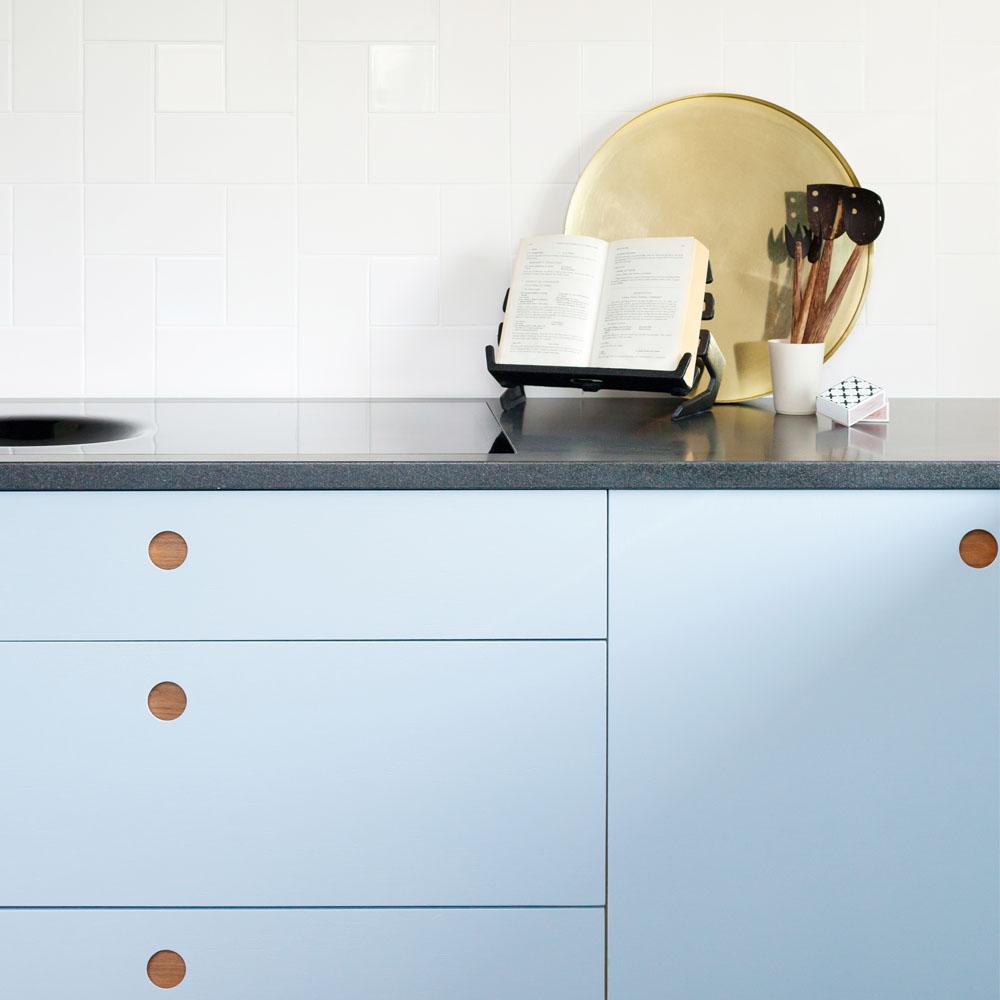 A modern kitchen with light blue cabinets, round wooden handles, a dark countertop, and white tiled backsplash. On the counter are a gold tray, an open cookbook on a stand, utensils in a white holder, and a sink.