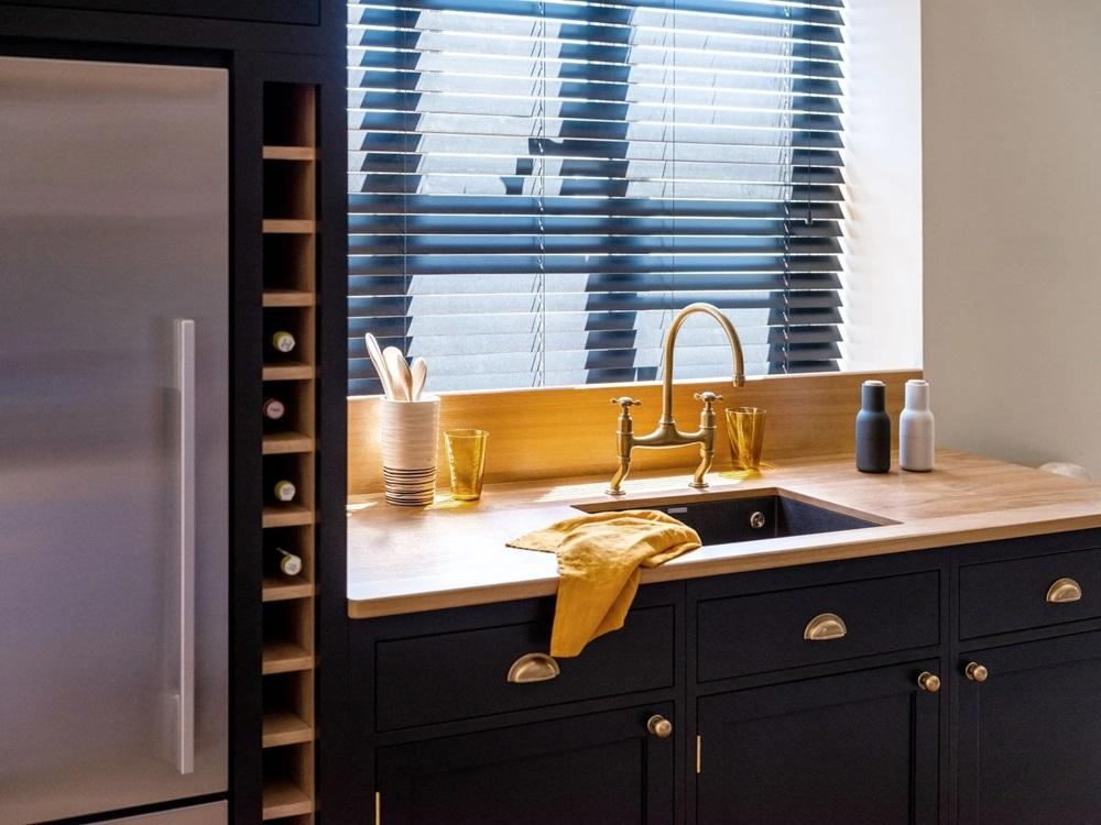 Black kitchen cabinets featuring brass shell handles, a wooden countertop, an undermount sink with a brass gooseneck faucet, and a built-in vertical wine rack next to a stainless steel refrigerator.