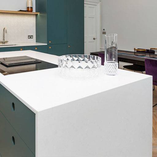 A modern kitchen with teal cabinets, a white countertop, a glass bowl, and glassware. In the background, a dining table with purple chairs and a wooden floor are visible.