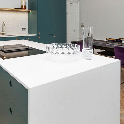 A modern kitchen with teal cabinets, a white countertop, a glass bowl, and glassware. In the background, a dining table with purple chairs and a wooden floor are visible.