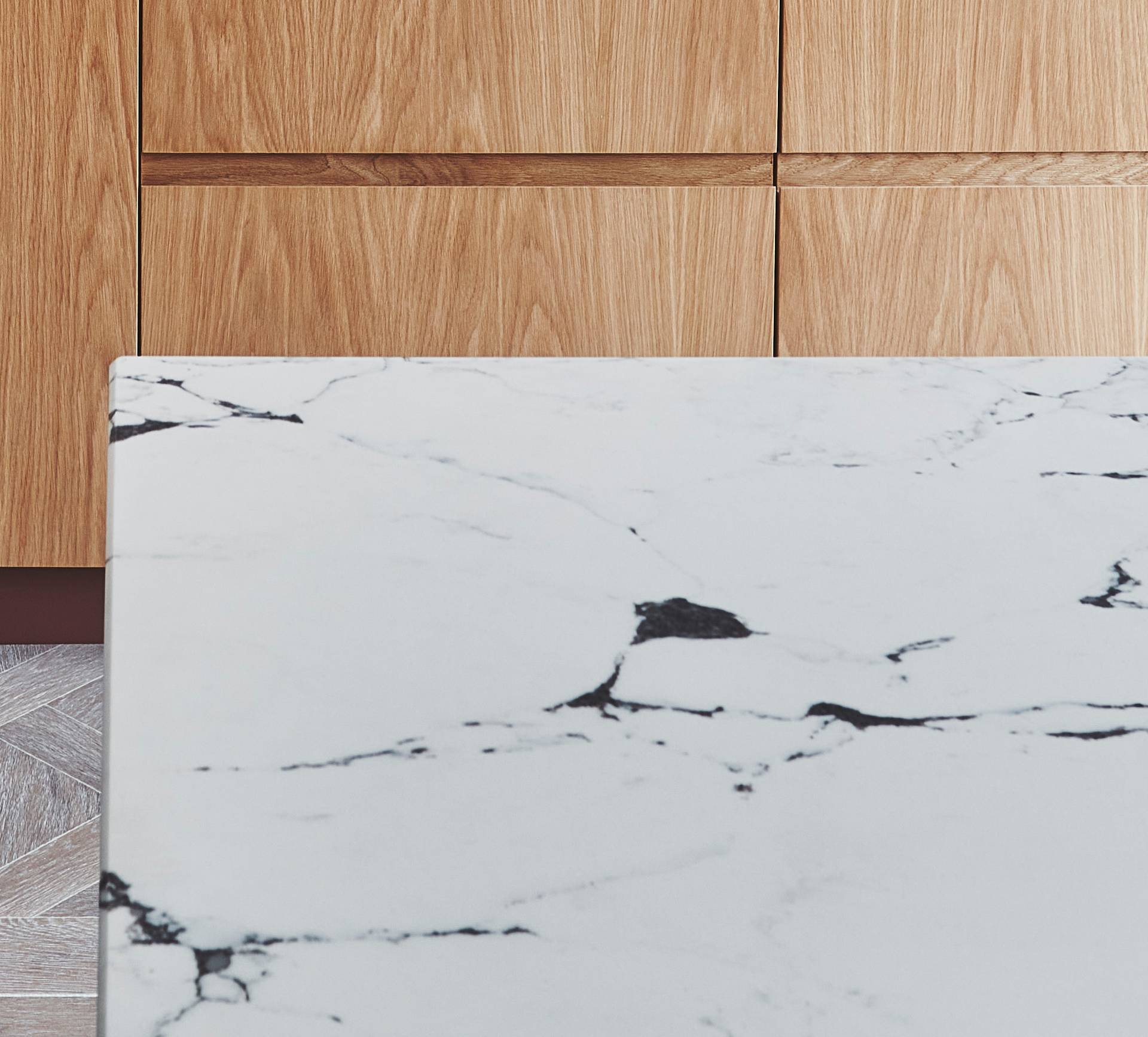Close-up of a kitchen counter with a white marble surface featuring dark veins. In the background, there are wooden cabinets with a light brown finish. The floor is partially visible, showing a gray and white tile pattern.
