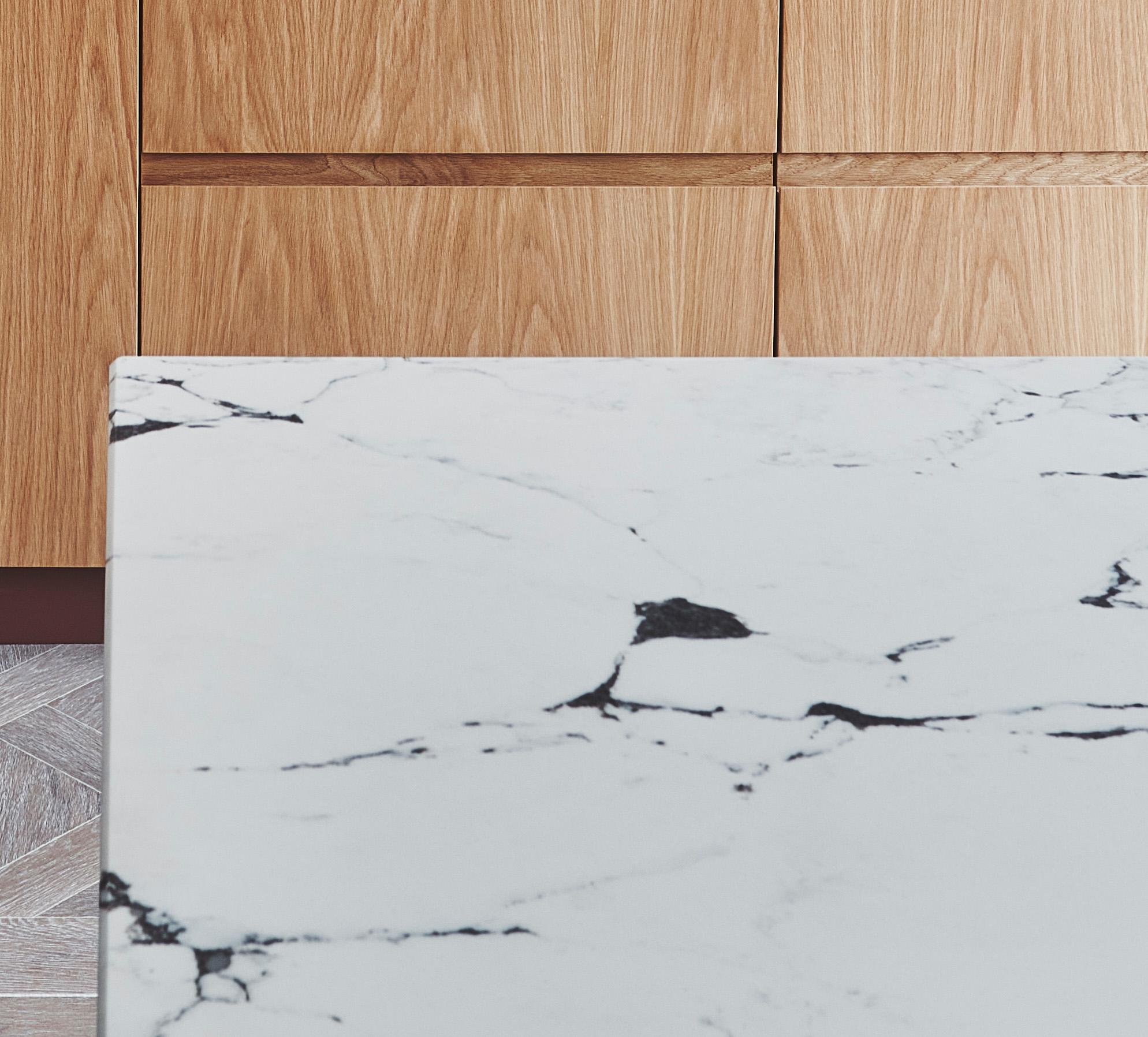 Close-up of a kitchen counter with a white marble surface featuring dark veins. In the background, there are wooden cabinets with a light brown finish. The floor is partially visible, showing a gray and white tile pattern.