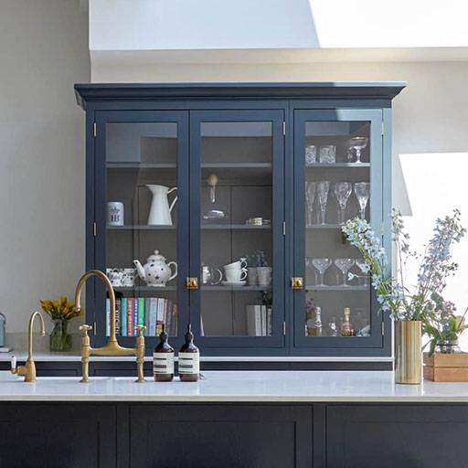 A modern kitchen with a dark blue glass-front cabinet displaying glassware, teapots, cups, and books. A white countertop features a brass faucet, soap bottles, and a gold vase with flowers.