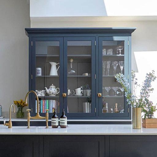 A modern kitchen with a dark blue glass-front cabinet displaying glassware, teapots, cups, and books. A white countertop features a brass faucet, soap bottles, and a gold vase with flowers.