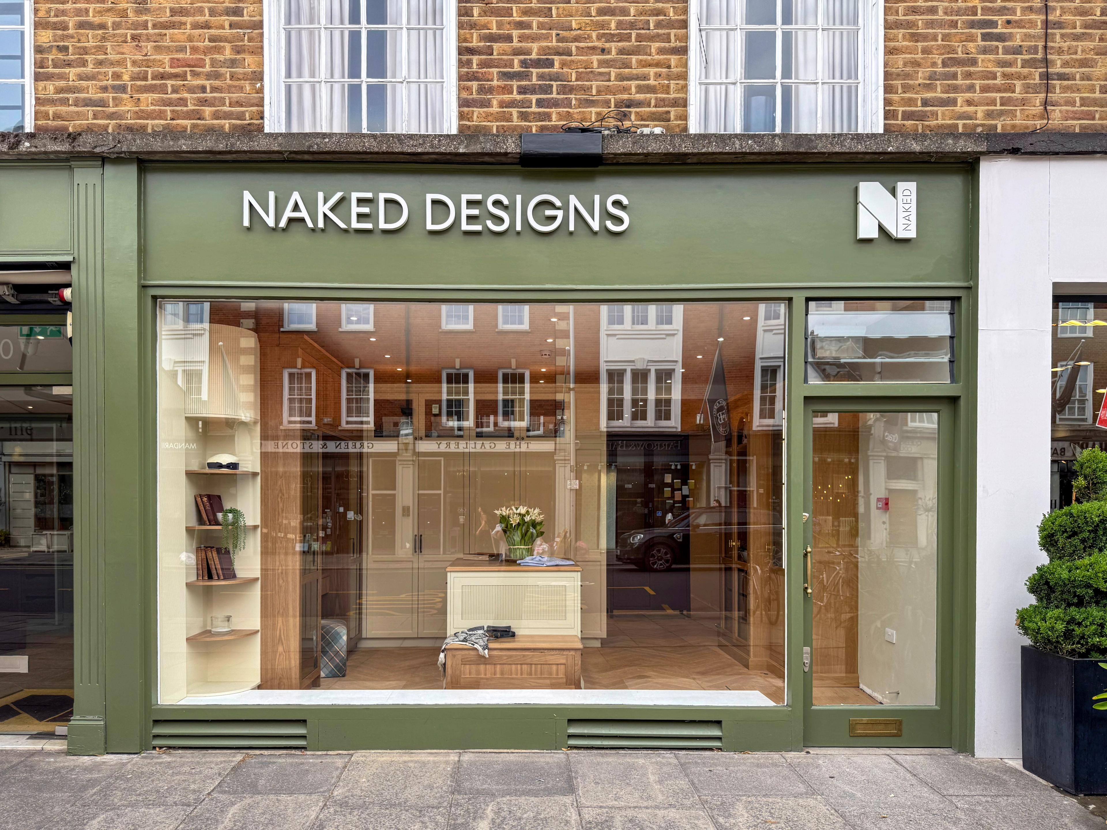 Street view of Naked Kitchens storefront with a green facade and glass door. An awning displays 'built for your life'. A small sign with the logo and name stands on the sidewalk. The interior shows a modern kitchen display.