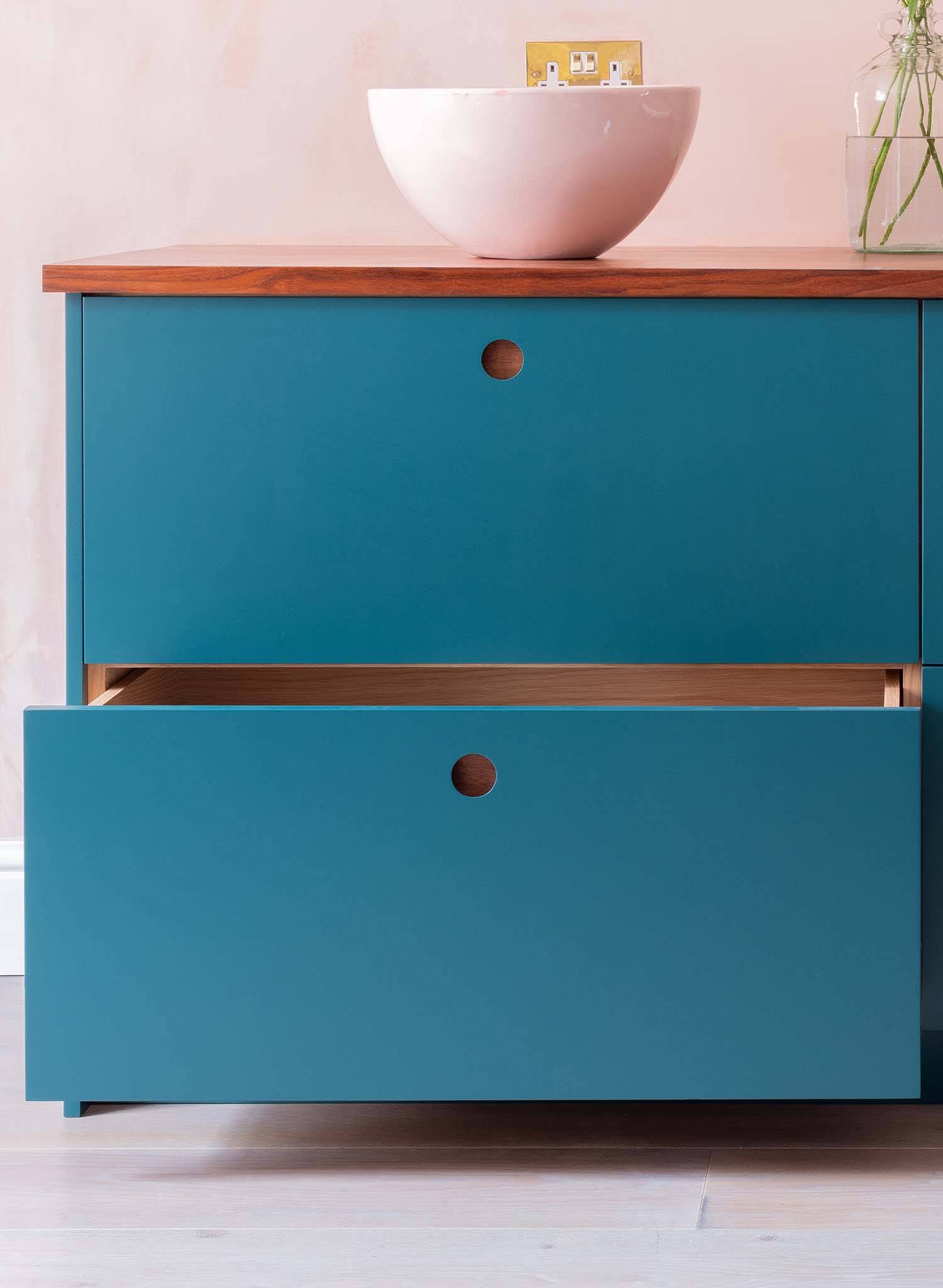 A modern blue cabinet with two drawers, the lower drawer partially open, stands on a light wood floor. A wooden countertop holds a white bowl and a glass vase with green stems.