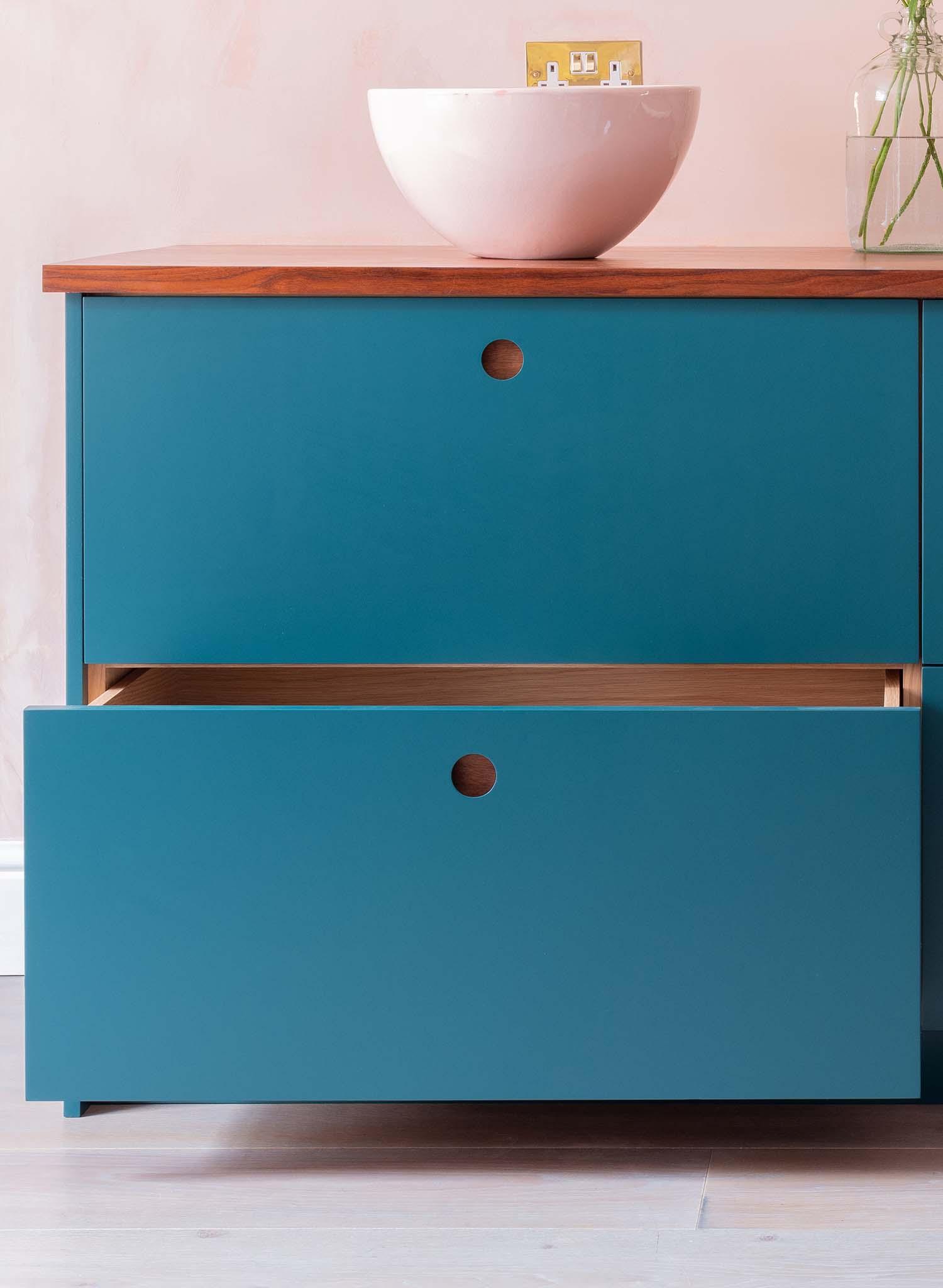 A modern blue cabinet with two drawers, the lower drawer partially open, stands on a light wood floor. A wooden countertop holds a white bowl and a glass vase with green stems.