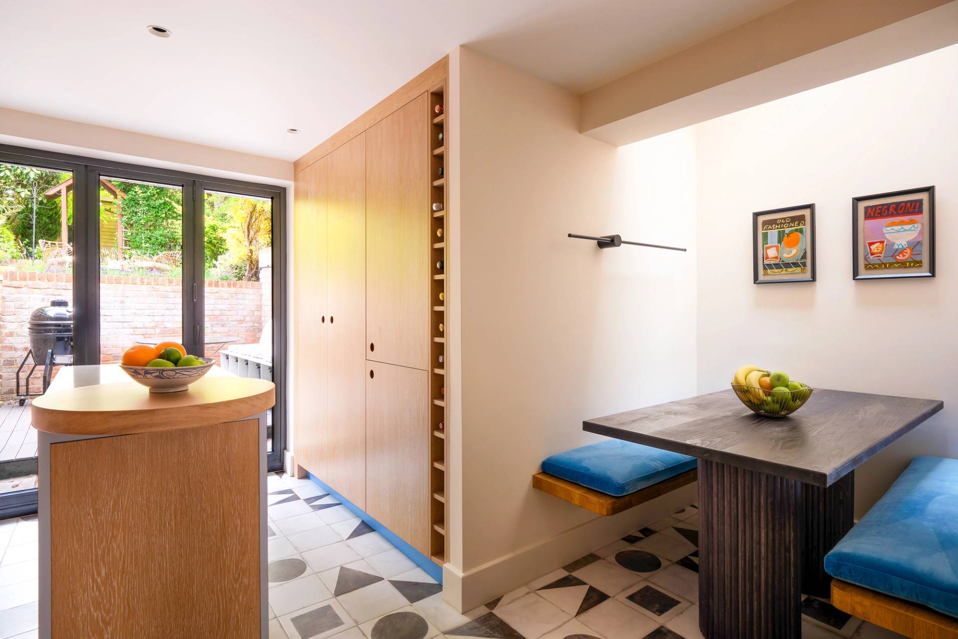 A modern kitchen with black-framed glass doors leading to a patio. The room features a small wooden dining table with blue cushioned benches, a fruit bowl on the table, and two framed posters on the wall. A tall wooden cabinet with wine storage is nearby