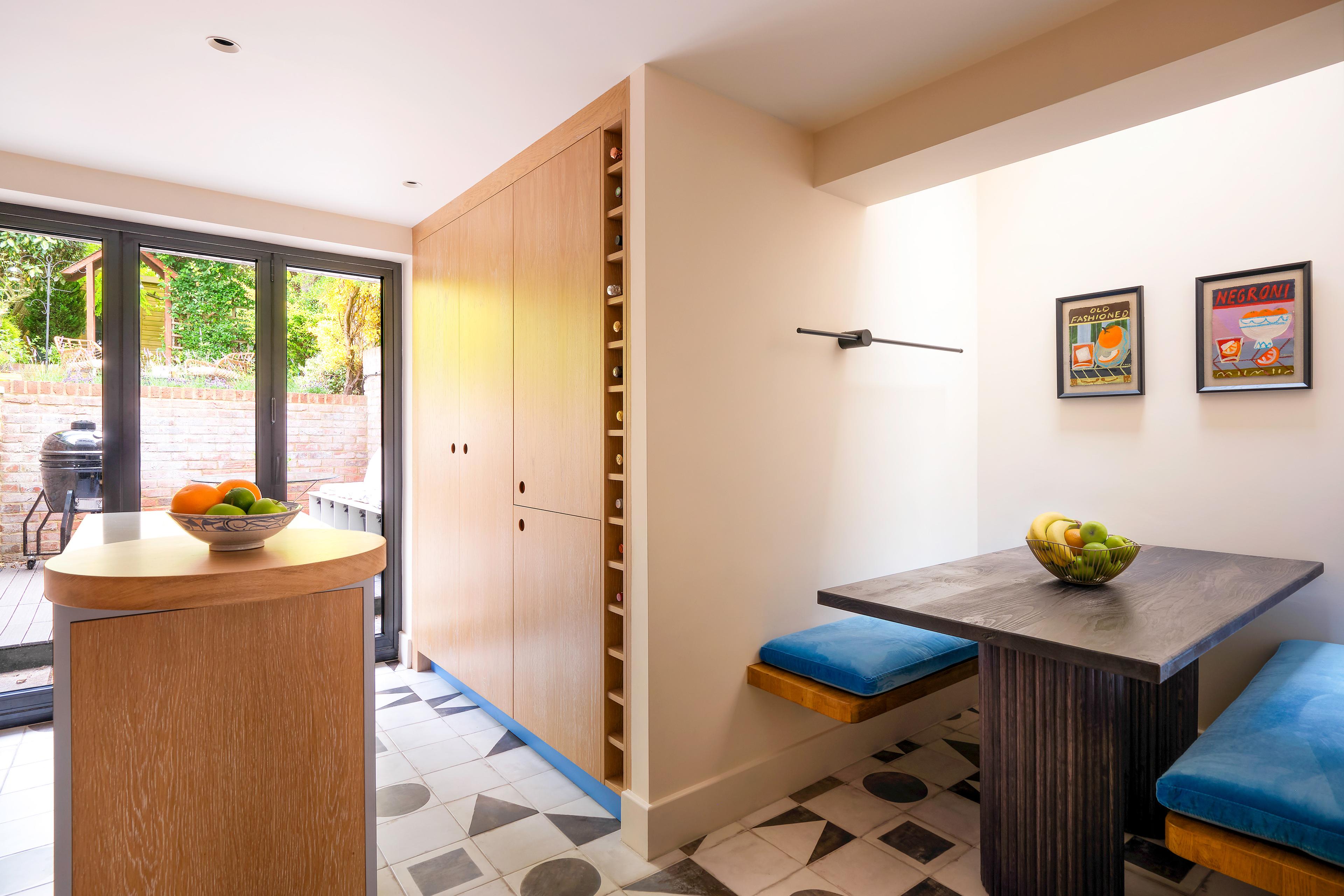 A modern kitchen with black-framed glass doors leading to a patio. The room features a small wooden dining table with blue cushioned benches, a fruit bowl on the table, and two framed posters on the wall. A tall wooden cabinet with wine storage is nearby