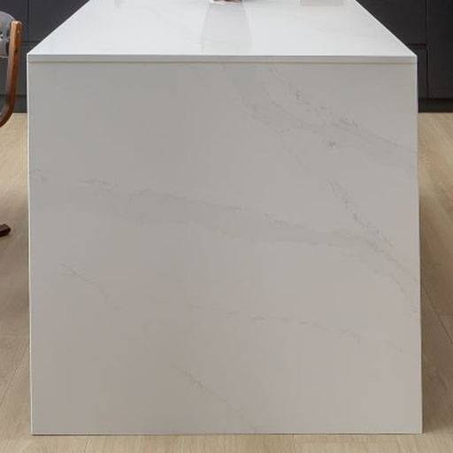 A minimalist kitchen island with a white, marble-look surface and subtle grey veining, resting on a light wood floor. Part of a wooden chair is visible on the left side.
