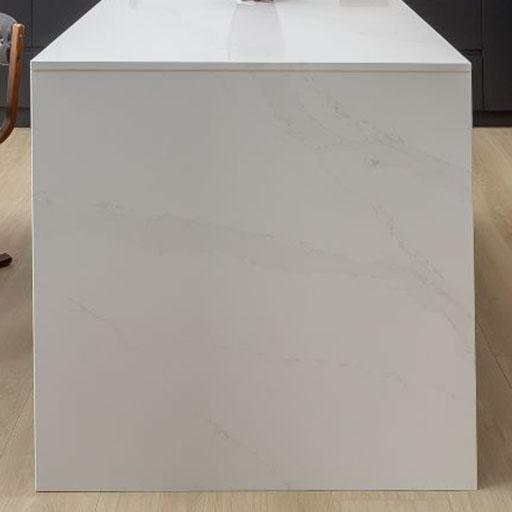 A minimalist kitchen island with a white, marble-look surface and subtle grey veining, resting on a light wood floor. Part of a wooden chair is visible on the left side.