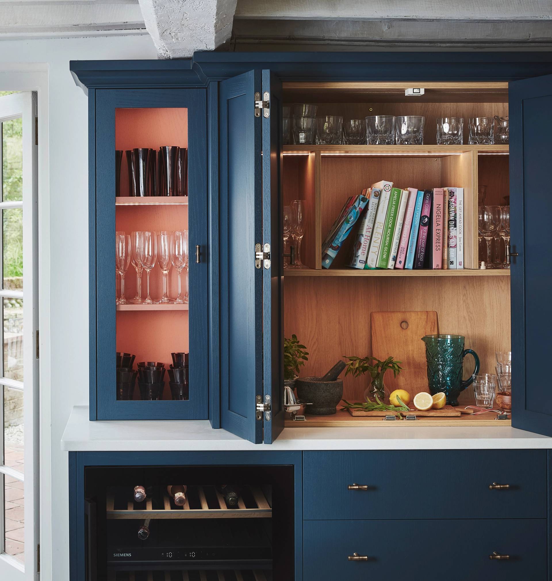 A blue kitchen cabinet with open doors displays glassware, books, and a cutting board with lemons, limes, and a knife on a white countertop. There’s also a wine rack and sunlight coming through a glass door nearby.