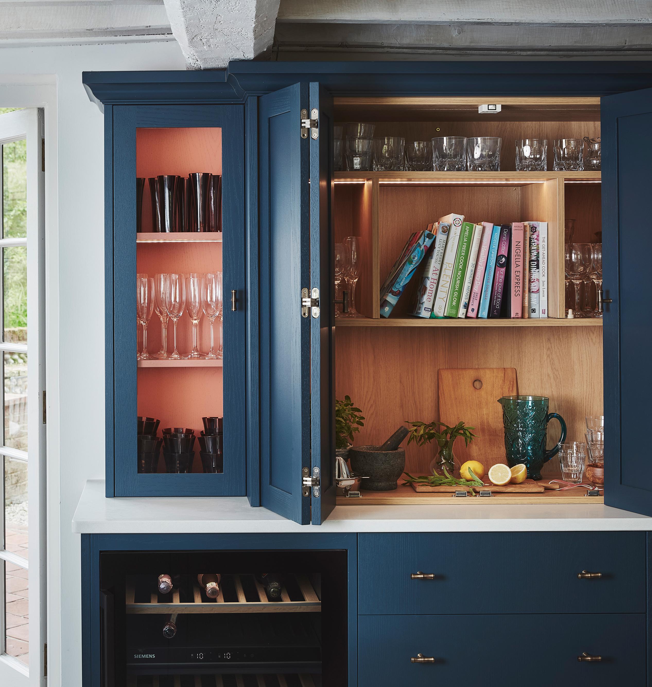 A blue kitchen cabinet with open doors displays glassware, books, and a cutting board with lemons, limes, and a knife on a white countertop. There’s also a wine rack and sunlight coming through a glass door nearby.
