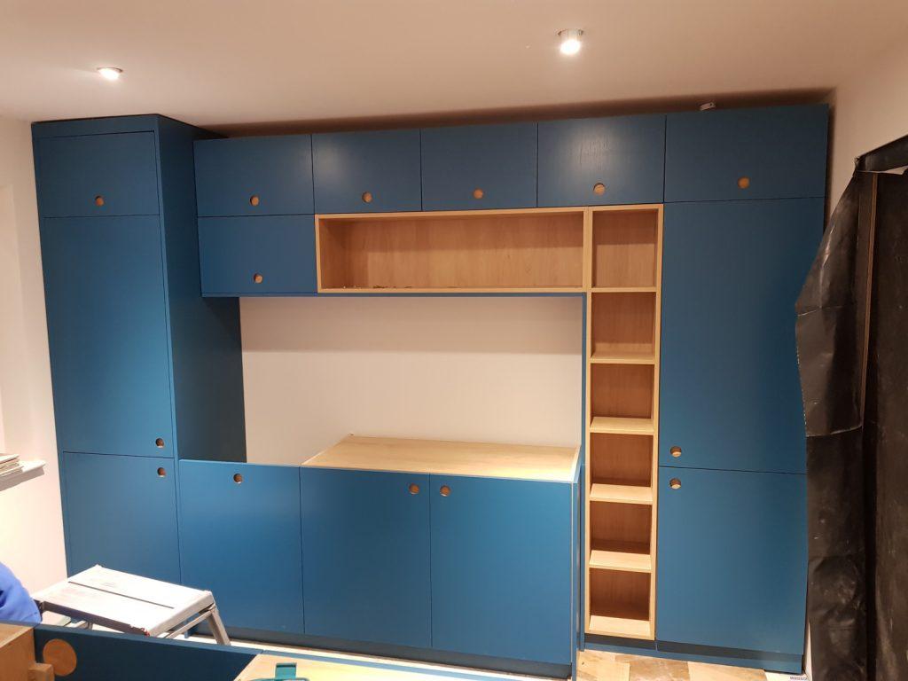 A wall of teal blue kitchen cabinetry featuring integrated natural wood shelving and round cutout handles during installation.