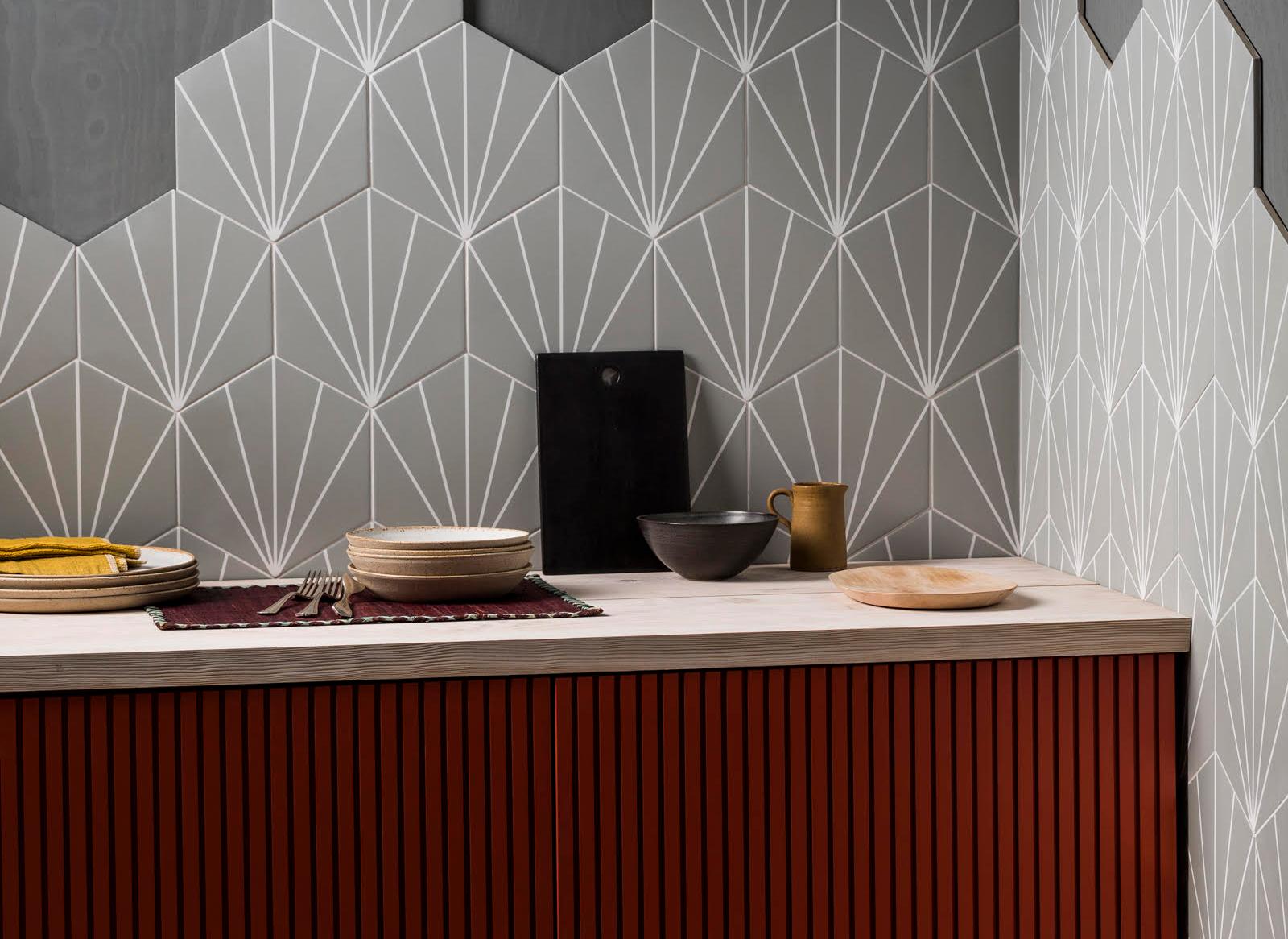 Kitchen with a modern design featuring geometric wall tiles in gray and white. A wooden countertop holds a cutting board, ceramic bowls, plates, and a mug. The countertop front has vertical slats in a rich reddish-brown tone.