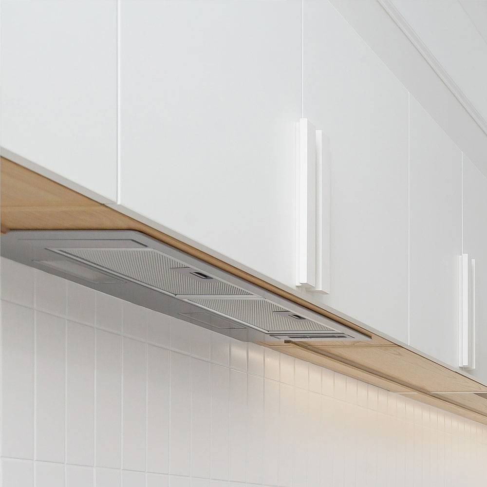 A close-up view of modern white kitchen cabinets with sleek handles, mounted above a tiled backsplash. A built-in stainless steel range hood is integrated underneath the cabinets.