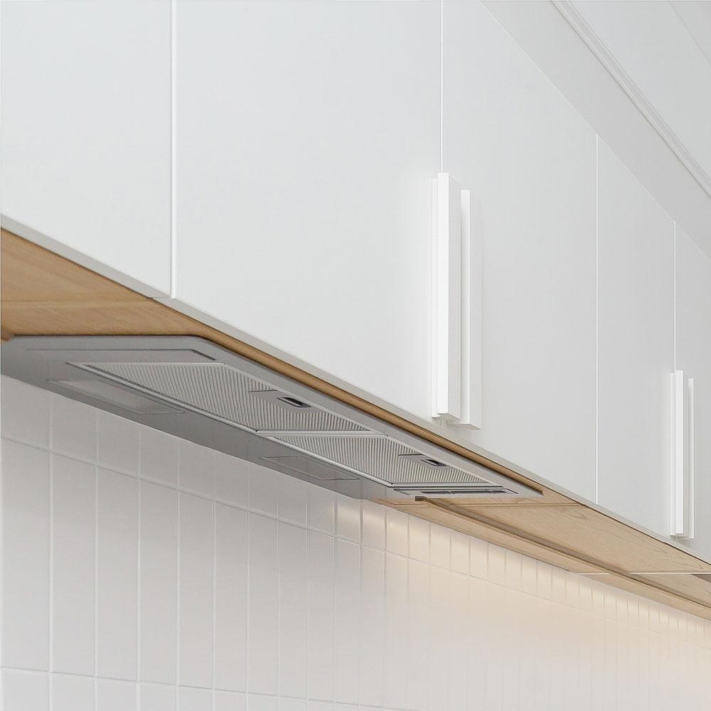 A close-up view of modern white kitchen cabinets with sleek handles, mounted above a tiled backsplash. A built-in stainless steel range hood is integrated underneath the cabinets.
